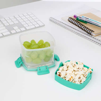Durchsichtige Snackbox mit grünen Trauben und Popcorn, auf einem Schreibtisch mit Bürobedarf.