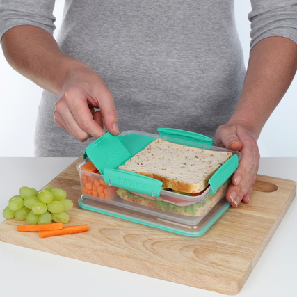 Hand, die eine sistema Lunchbox öffnet, auf einem Holzbrett mit Trauben und Karotten daneben.