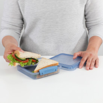 Person hält eine Sandwichbox mit einem halbierten Sandwich auf einem Tisch.