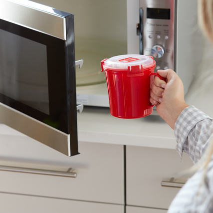 Eine Person hält eine rote Mikrowellensuppe-Tasse vor einem geöffneten Mikrowellenofen.