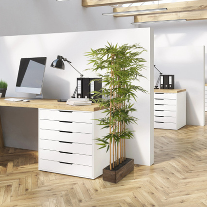 Künstliche Bambuspflanze in braunem Holzbehälter mit Zementfuß in einem Büroambiente.