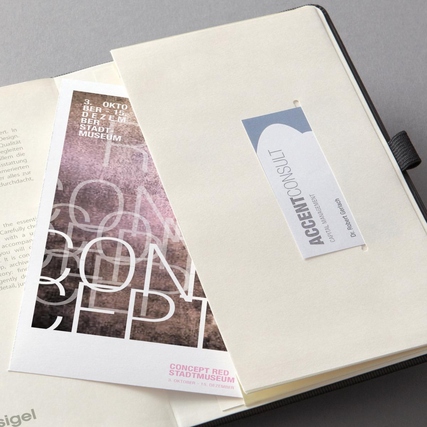 Offene Buchkalenderseite mit eingelegtem Flyer und Visitenkarte, auf einer neutralen Oberfläche.