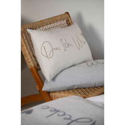 Hellgrauer Kissenbezug mit Aufschrift "Du. Ich. Wir." auf einem Rattanstuhl.