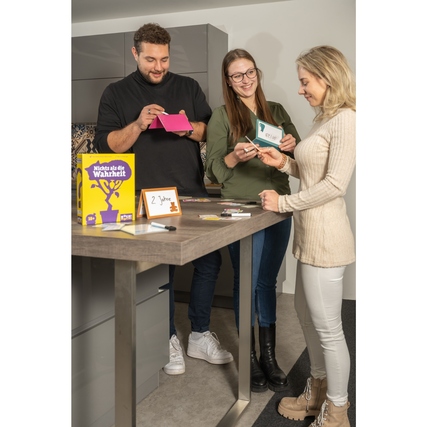 Drei Personen stehen an einem Tisch und spielen das Gesellschaftsspiel 'Nichts als die Wahrheit'.