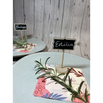 Zwei beschriftete Holzschilder auf Tellern mit Servietten und frischen Kräutern als Dekoration.