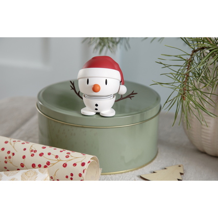 Dekorationsfigur Schneemann mit Weihnachtsmütze auf einer grünen Dose, umgeben von Geschenkpapier.