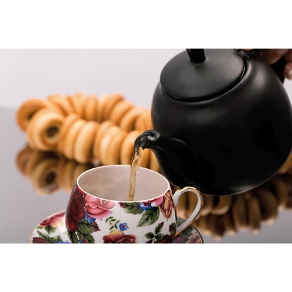 Eine Teekanne gießt heißen Tee in eine dekorative Tasse, im Hintergrund sind Gebäcke sichtbar.