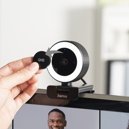 Eine Hand hält eine Linse vor einer Webcam mit Ringlicht, die auf einem Monitor montiert ist.