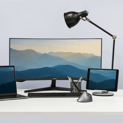 Ergonomische PC-Maus in einem Büroumfeld mit Monitoren, Laptop und Tischlampe.