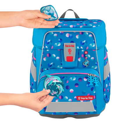 Zwei Hände befestigen farbige Anhänger an einem blauen Schulranzen mit bunten Punkten.