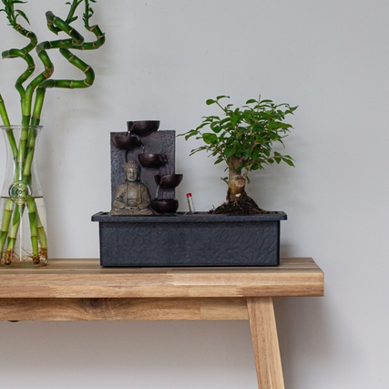 Bonsai-Baum in Pflanzgefäß mit Wasserfall und Buddha auf einem Tisch.