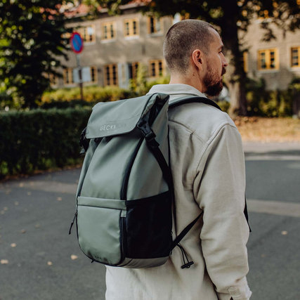 Rucksack in Lichtgrün auf einem Träger, Hintergrund mit Gebäuden.