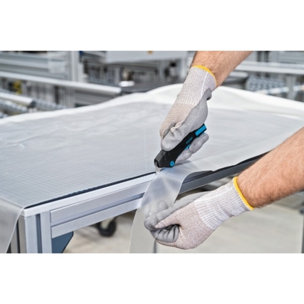 Person mit Handschuhen nutzt ein Sicherheitsmesser zum Schneiden eines Materials auf einem Tisch.