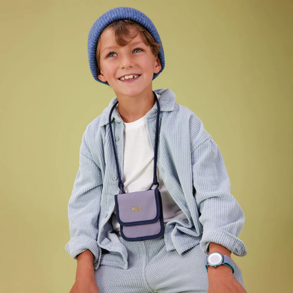 Ein Kind trägt einen blauen Brustbeutel in einem modischen Outfit vor einem neutralen Hintergrund.