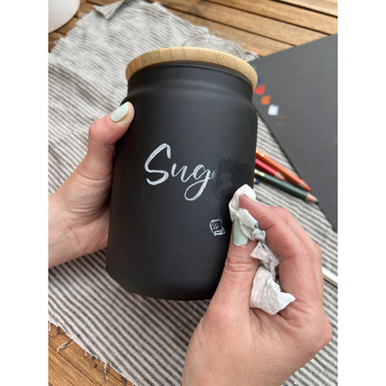 Buntstift schreibt auf einem Glasbehälter, während eine Hand diesen mit einem Tuch abwischt.