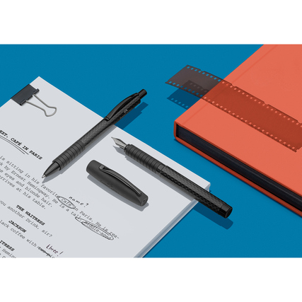 Füllfederhalter und Stift auf einem Blatt Papier, umgeben von Zubehör und einem Notizbuch.