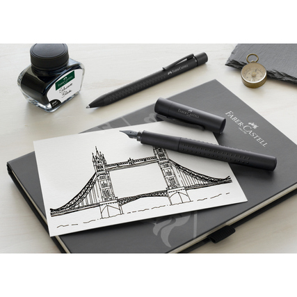 Füllfederhalter in Schwarz neben Tinte, Skizze und Notizbuch auf einem Tisch.