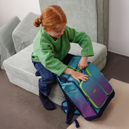 Schulranzen in türkis-violett mit neonfarbenen Akzenten, neben einem Kind auf einem Sofa.