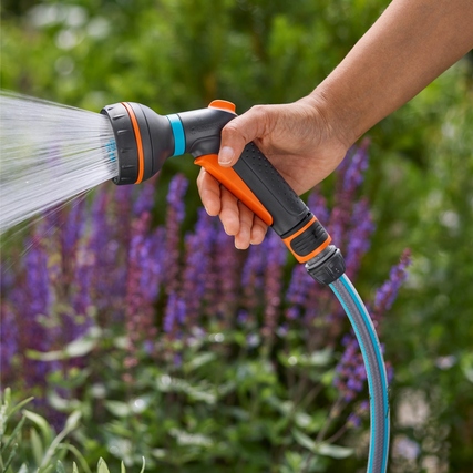 Multifunktionsbrause in Hand mit sprühendem Wasser, umgeben von grünen Pflanzen.