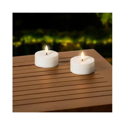 Zwei weiße LED-Kerzen auf einem Holztisch, umgeben von einer unscharfen Gartenlandschaft.