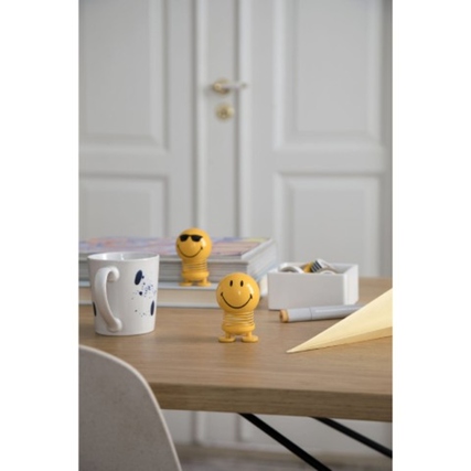 Dekorationsfigur Smiley in Gelb auf einem Tisch, umgeben von Büroartikeln und einem Becher.