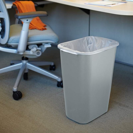 Abfallbehälter in Grau steht unter einem Schreibtisch auf einem Teppichboden.
